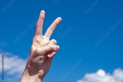 Two fingers of a caucasian woman hand showing victory or peace gesture on blue sky background. Copy space