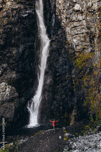 noise effect  selective focus  miniature tourist dressed in a bright colored raincoat that is contrastingly visible on a large scale background of a cliff with a waterfall