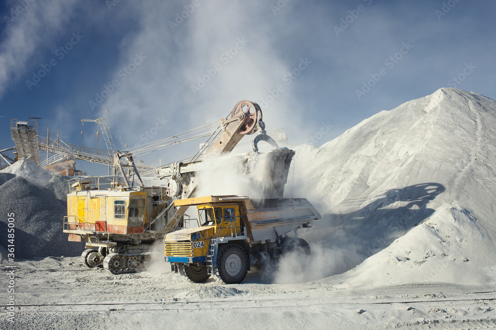 Mining excavator and heavy mining truck during loading in a cloud of dust on the background of the equipment of the mining enterprise in sunny weather. Mining industry. Mining excavator and heavy mining truck during loading in a cloud of dust on the background of the equipment of the mining enterprise in sunny weather. Mining industry.