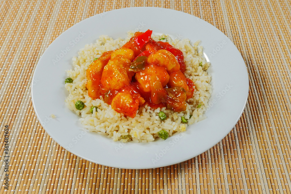 Serving of Chinese style sweet and sour chicken with rice on a white plate and bamboo table. Serving of Chinese style sweet and sour chicken with rice on a white plate and bamboo table.