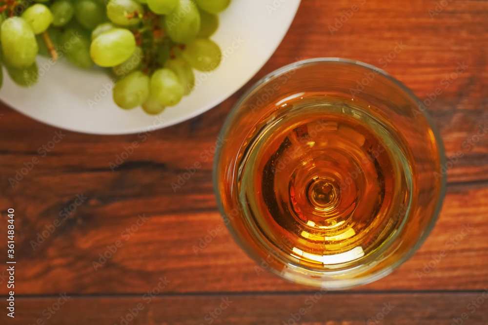 Top view on glass of white wine on dark wood table in focus, Cluster of grapes on a white plate out of focus. Top view on glass of white wine on dark wood table in focus, Cluster of grapes on a white plate out of focus.