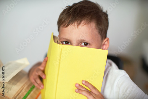 smart boy with glasses hides his face behind a book with a blue cover