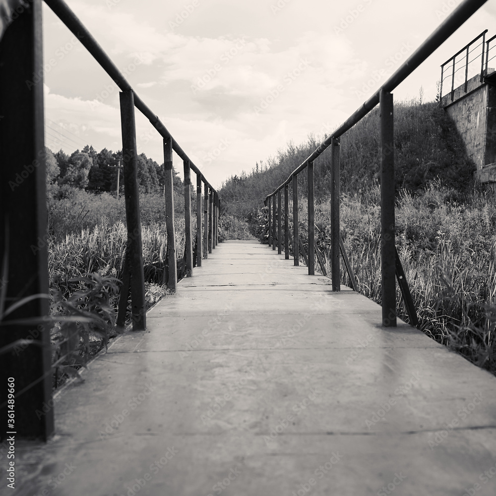 Fototapeta premium Black and white square photo of an old iron bridge on a background of beautiful greenery and trees 