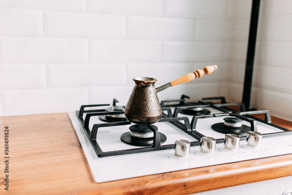 Coffee brewing at home in jezve on gas oven. Coffee cups to go on the wooden table. White kitchen interior. Wooden complete kitchen with gas oven