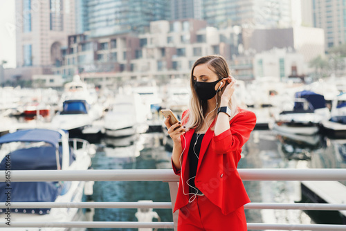 Young european woman with blond hair dressed in stylish red business suit and wearing black face mask standing in front of yachts and skyscrapers in city business center and making a call  © VeronikaSmirnaya