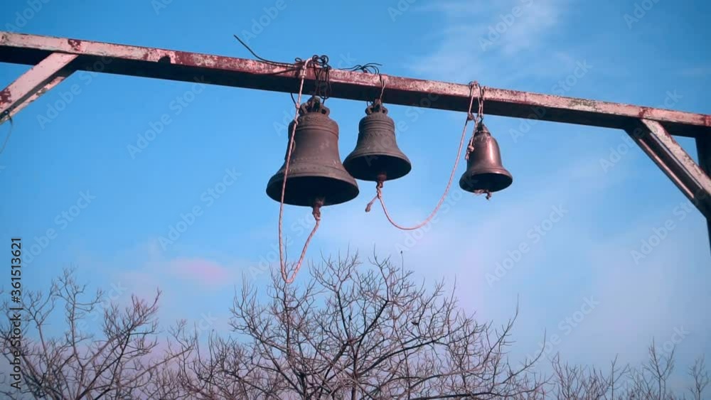 Bells on a wooden crossbar. Church bells. In the background are trees and blue sky. Bells on a wooden crossbar. Church bells. In the background are trees and blue sky.