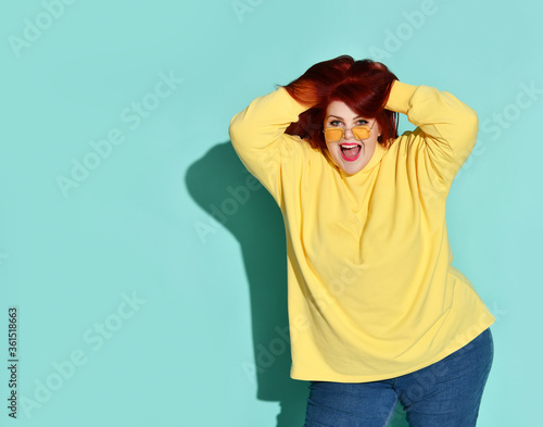 Cute young woman with overweight smiling joyfully running fingers through her long ginger hair. Three quarter length shot isolated on blue © FAB.1