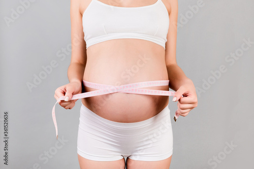 Close up of pregnant woman in white underwear measuring her growing tummy with a tape measure at gray background. Check a baby development. Copy space