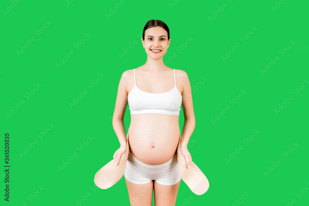 Portrait of pregnant woman in underwear dressing maternity belt against pain in the back at green background with copy space. Orthopedic abdominal support belt concept Portrait of pregnant woman in underwear dressing maternity belt against pain in the back at green background with copy space. Orthopedic abdominal support belt concept
