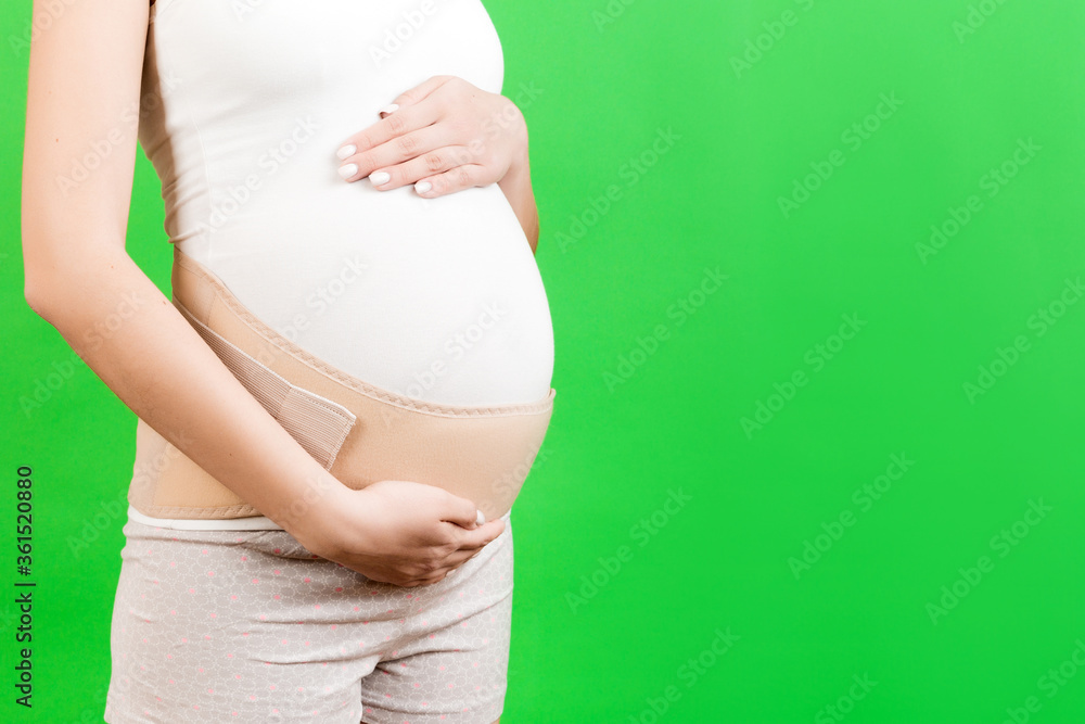 Close up of pregnant woman dressed in orthopedic corset for reducing pain in the back at green background with copy space. Orthopedic abdominal support belt concept Close up of pregnant woman dressed in orthopedic corset for reducing pain in the back at green background with copy space. Orthopedic abdominal support belt concept