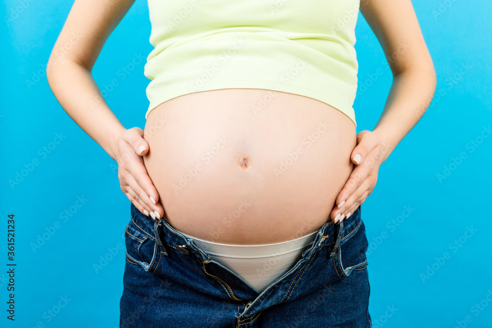 Close up of pregnant woman in opened jeans caressing her baby bump at colorful background with copy space. Waiting for a baby concept Close up of pregnant woman in opened jeans caressing her baby bump at colorful background with copy space. Waiting for a baby concept