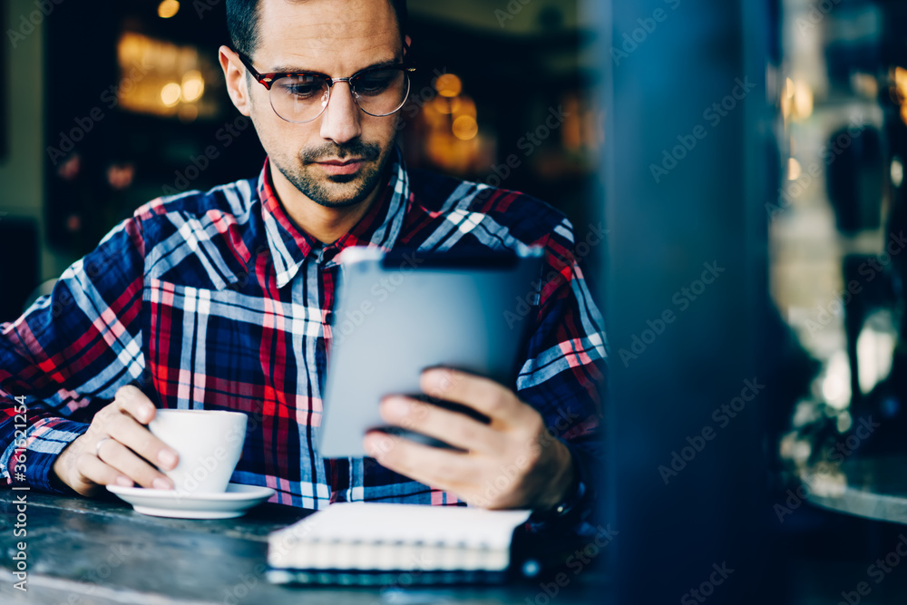Successful young bearded man in eyewear reading news on websites via high speed 4G internet connected to digital tablet while sitting indoors in coffee shop and enjoying tasty drink in free time Successful young bearded man in eyewear reading news on websites via high speed 4G internet connected to digital tablet while sitting indoors in coffee shop and enjoying tasty drink in free time