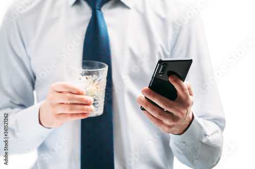 A guy in a white shirt and blue tie on a white background. A man looks at the phone screen and holds a glass with a cocktail.