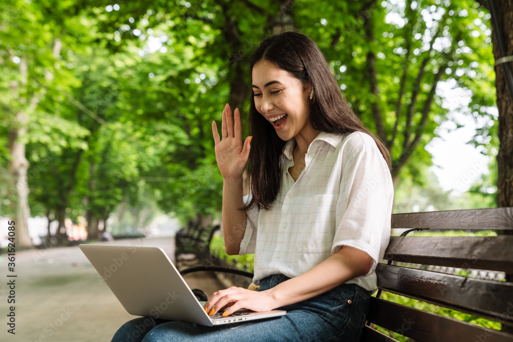 Photo of asian woman using laptop and waving hand while sitting on bench Photo of asian woman using laptop and waving hand while sitting on bench