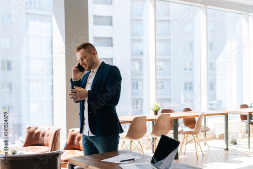 Handsome young businessman wearing fashion suit is talking on mobile phone in modern office room near wooden desk on background of large window. Concept of office working.
