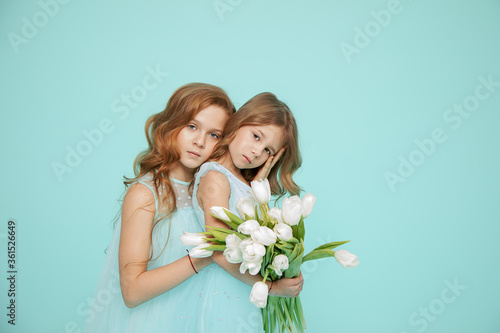 Two beautiful girls with curly locks of hair in dresses are embracing on blue background. Photo. White Tulip. Hair salon. Psychology of relationships. Look at camera. Friendship, love, family, touch © Татьяна Волкова
