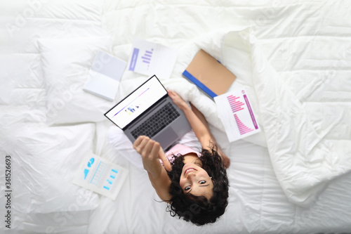 Top view of smiling happy female showing thumbs up. Young attractive woman working from home. Sweet lady with laptop and papers in bed. Remote job and freelance concept