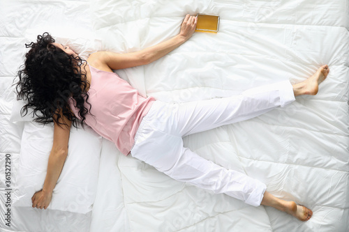 Top view of drunk female sleeping on white bedding holding alcohol bottle. Young woman resting after fun night party. Lady wearing light pants and pink shirt for sleep. Hungover and tired concept photo