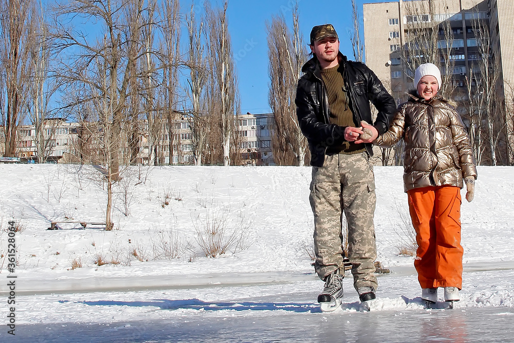 a young man on skates holds a little girl on skates on a ice skating rink in winter day