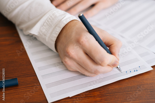 Close up of hands of unrecognizable man writing musical notes with pencil on sheet music, sitting at the wooden desk in modern room.