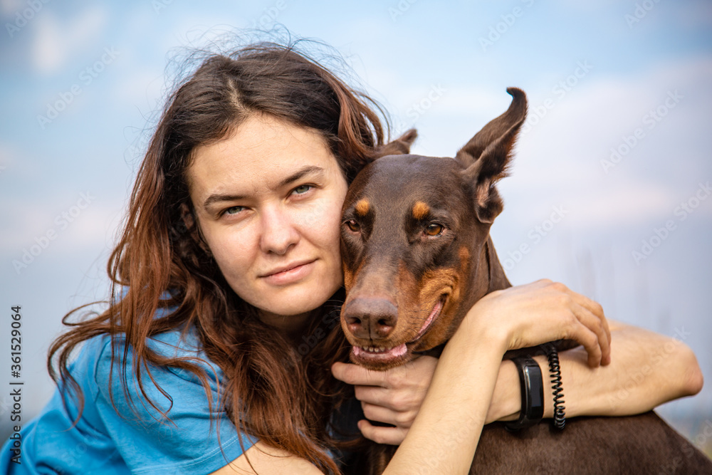 Girl in a blue T-shirt hugs a Doberman Dobermann dog. Portrait against the sky. Horizontal orientation.