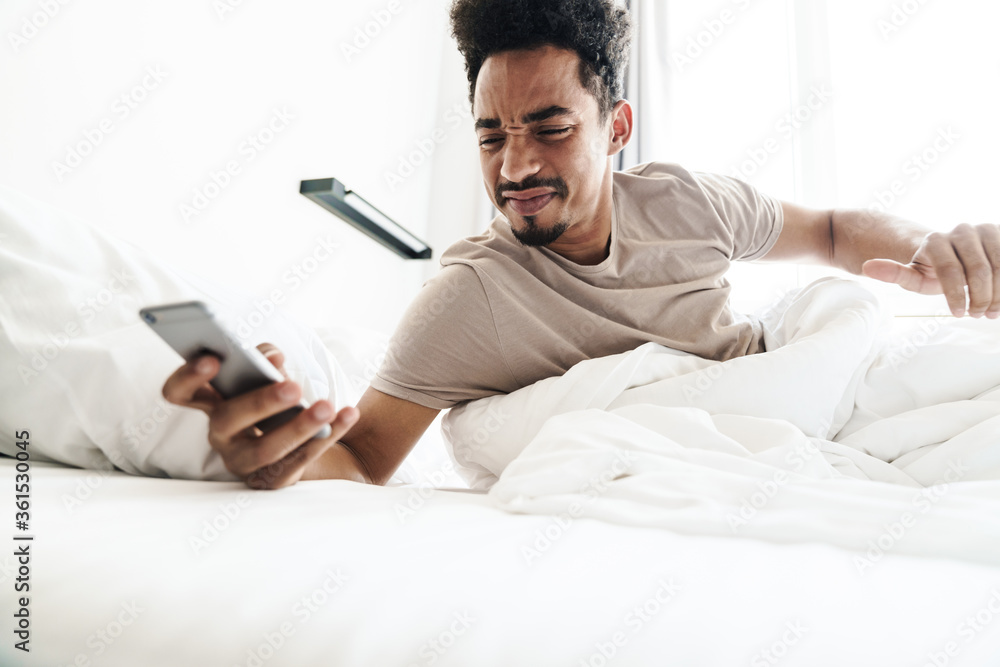 Fototapeta premium Photo of displeased african american man using mobile phone in white bed