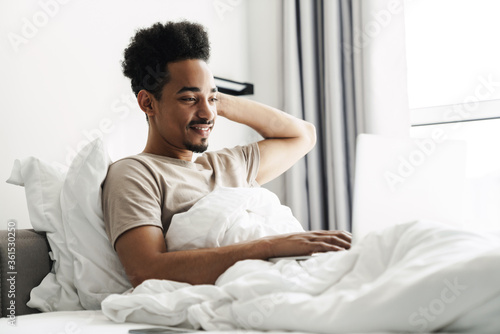 Photo of african american man working with laptop while lying in bed