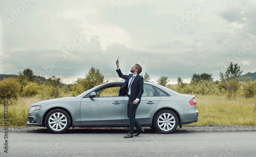Businessman walking around car on roadside in country area holding phone looking for mobile network