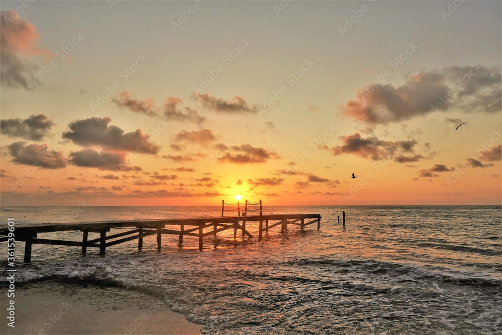 Beautiful sunrise over the Caribbean. The sun rose slightly above the horizon and painted the sky in scarlet and gold colors. A wooden path goes over the sea. In the sky are picturesque clouds.