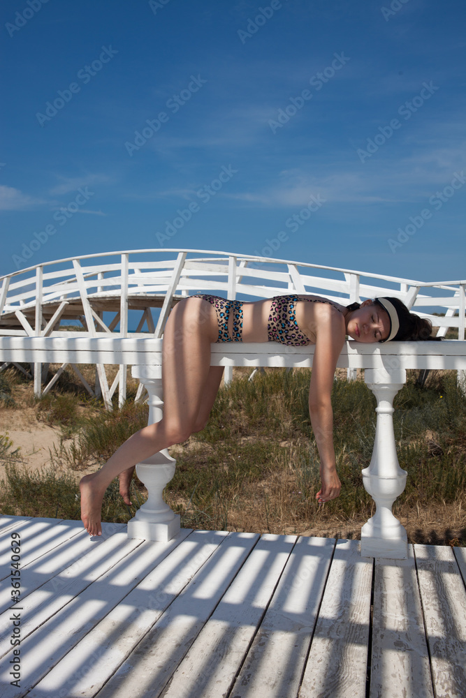 Fototapeta premium A young girl in a bathing suit is lying on the railing. Very tired. Funny pose of the model. Beach holiday.