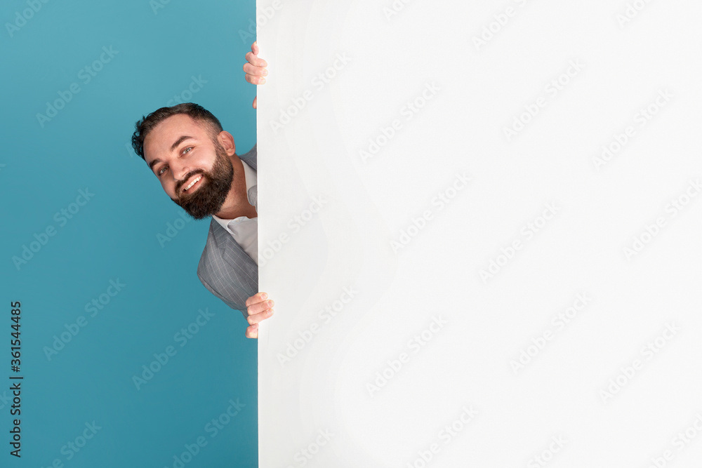 Smiling entrepreneur hiding behind blank poster with space for text or design on blue background Smiling entrepreneur hiding behind blank poster with space for text or design on blue background