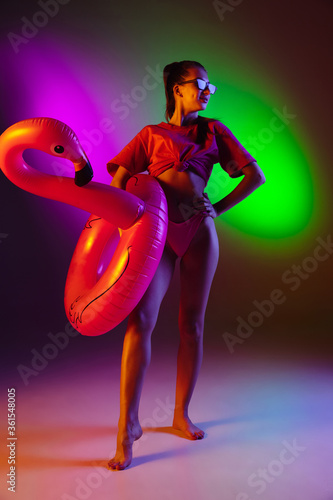 Seductive young girl' portrait isolated on bicolored neon studio background in neon. Fit sportive woman in bodysuit with rubber flamingo. Facial expression, summer, weekend, beauty, resort concept.