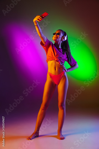 Seductive young girl' portrait isolated on bicolored neon studio background in neon. Fit sportive woman in bodysuit taking selfie. Facial expression, summer, weekend, beauty, resort concept. Vacations