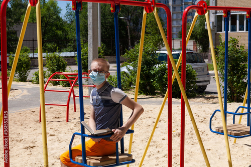 A boy in a protective mask on the Playground reading a book on a Sunny summer day. А new normal, new reality.
