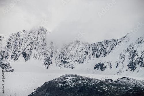 Black mountains with white snow on tops and glacier. Dramatic landscape with snowy mountains under cloudy gray sky in grayscale. Atmospheric alpine scenery with snow rocky mountains among low clouds.