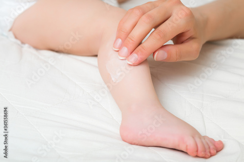 Young mother hand applying white moisturizing cream on baby leg. Care about children clean and soft body skin. Closeup.