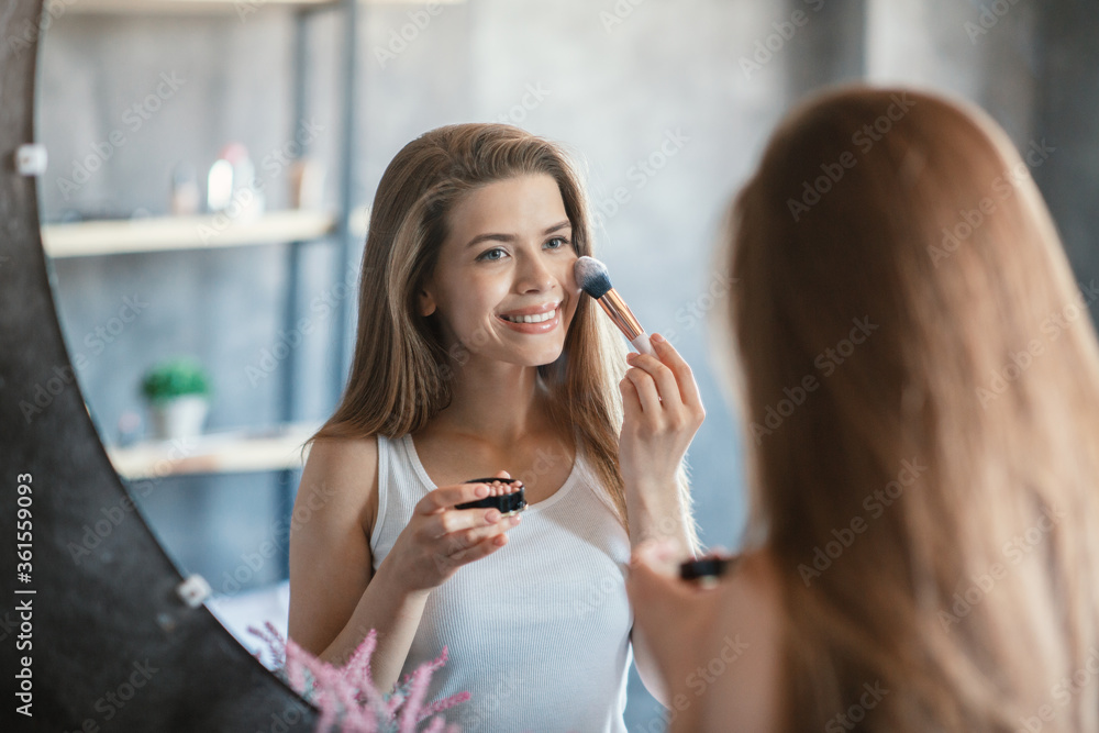 Pretty young woman applying blusher with cosmetic brush in front of mirror at bathroom Pretty young woman applying blusher with cosmetic brush in front of mirror at bathroom