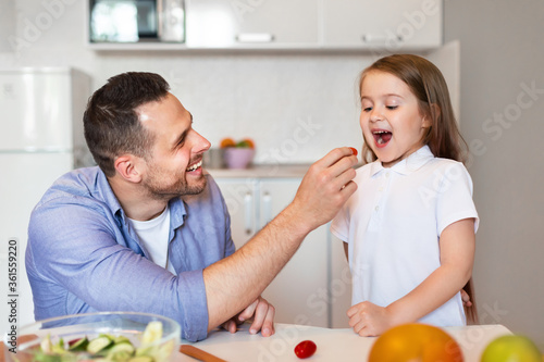 Father Feeding Little Daughter Preparing Dinner In Kitchen At Home