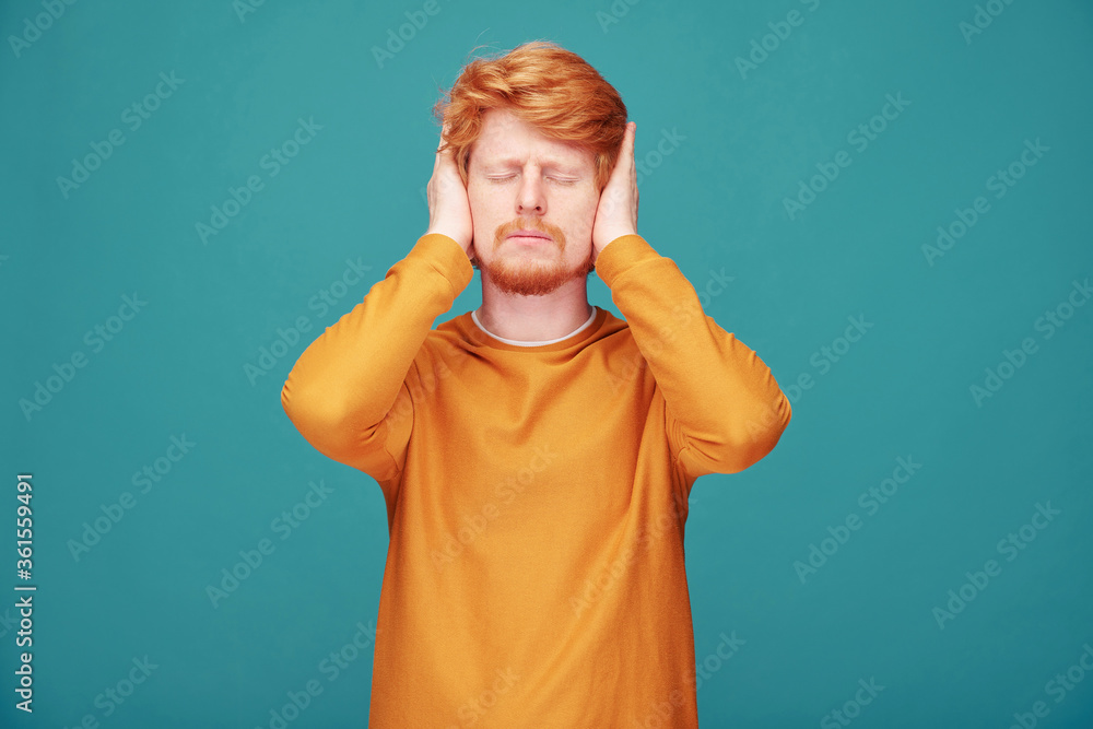 Annoyed young redhead man with beard keeping eyes closed and covering ears with hands against blue background Annoyed young redhead man with beard keeping eyes closed and covering ears with hands against blue background