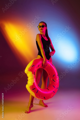 Seductive young girl' portrait on bicolored neon studio background in neon. Fit sportive woman in bodysuit with rubber donut. Facial expression, summer, weekend, beauty, resort concept. Vacations.