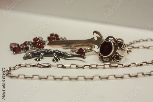 Silver pendant in the shape of a sword on an anchor chain, silver set of earrings and rings with natural red garnet stone, pendant in the shape of a lizard with transparent zircon on a white backgroun