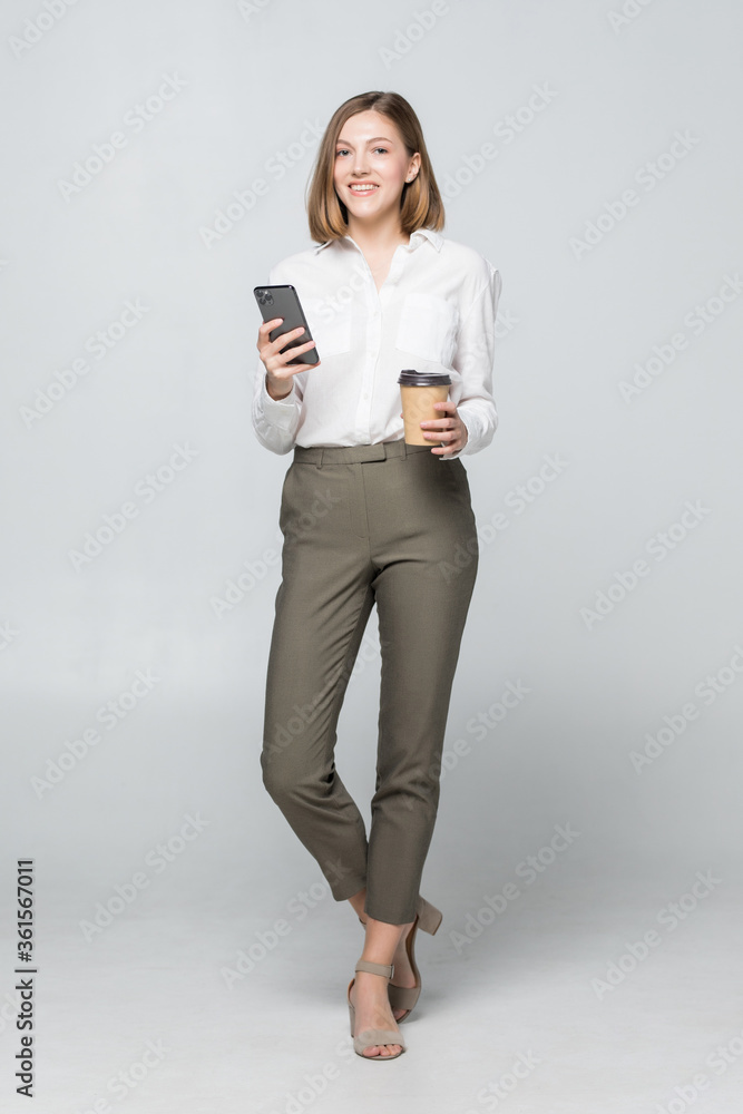Pretty business woman in formal wear standing and using cell phone with takeaway coffee in hand isolated over gray background Pretty business woman in formal wear standing and using cell phone with takeaway coffee in hand isolated over gray background