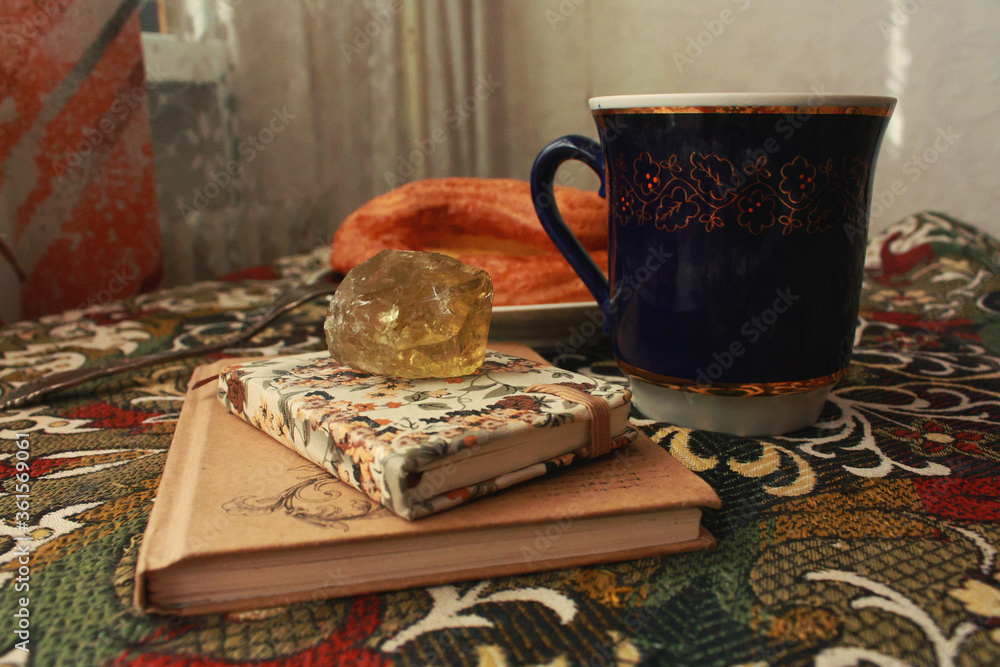 Fototapeta premium Composition of banana puff on a plate, a blue cup of tea, a fork, two notebooks and a citrine stone on a multi-colored tablecloth.