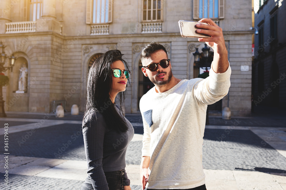 Stylish girl and her handsome friend doing selfie against the background the historic building. Young man holding metallic color phone. The sun is shining so brightly that couple put on sunglasses