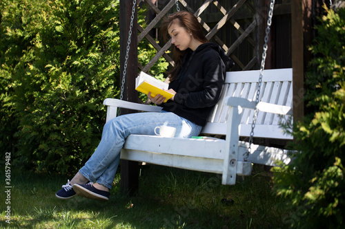Attractive brunette with long hair dressed in black hoodie sitting on white swing bench reading book with cup of coffee. Relaxed vacation concept. Distance Education Concept