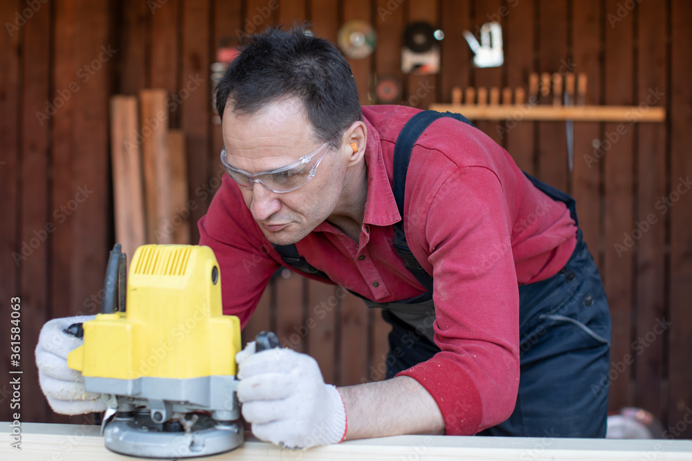 Handsome 40s carpenter works on wooden workpiece with milling tool at cottage workshop close up. Processing of Natural Wooden beam with milling woodworking machine. jack of all trades concept Handsome 40s carpenter works on wooden workpiece with milling tool at cottage workshop close up. Processing of Natural Wooden beam with milling woodworking machine. jack of all trades concept