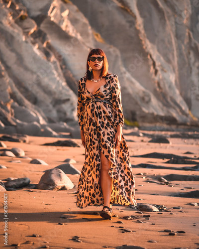 Summer street style on a beach next to rocks of a young brunette caucasian woman in a leopard dress. Sakoneta beach in the town of Deba, Basque Country. Walking the beautiful landscape