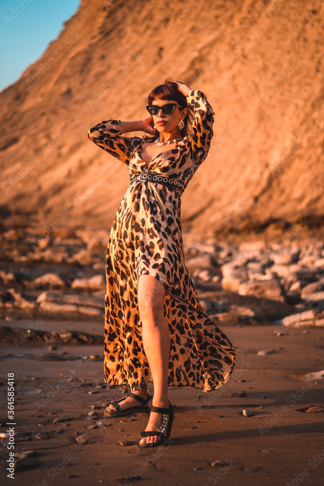 Summer lifestyle on a beach next to rocks of a young brunette Caucasian woman in a leopard dress by the sea. Sakoneta beach in the town of Deba, Basque Country. Summer lifestyle on a beach next to rocks of a young brunette Caucasian woman in a leopard dress by the sea. Sakoneta beach in the town of Deba, Basque Country.