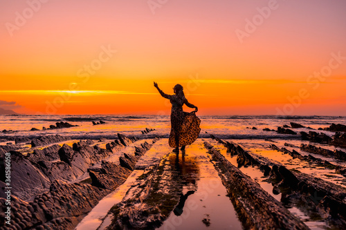 Lifestyle with sunset  a silhouette of a young girl in a dress on the beach of Sakoneta in the village of Deba  Gipuzkoa. Basque Country. Young girl holding dress in water next to rocks