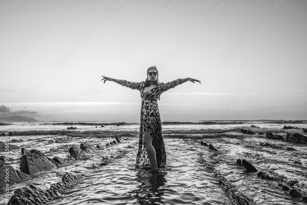Lifestyle, a young brunette woman in leopard dress on a sunset at the coast with sunglasses. Black and white photo of young girl in the water next to the rocks
